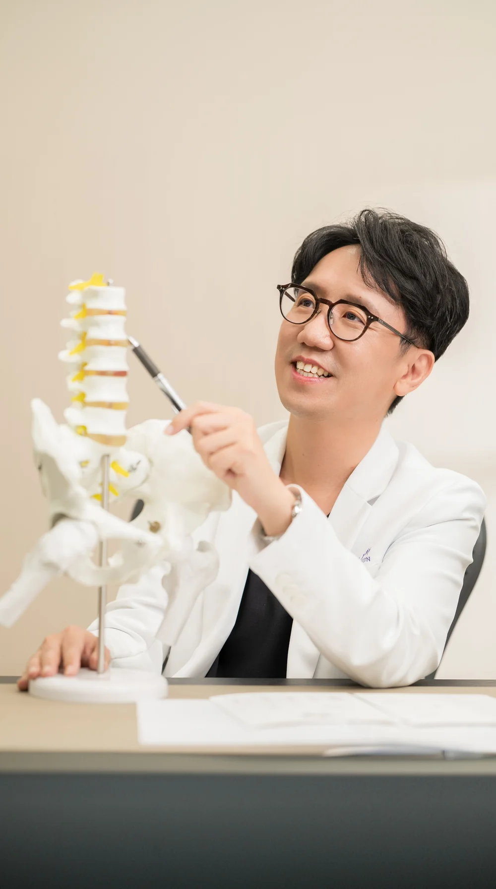 邱柏鈞院長身穿白袍，微笑手持筆指向脊椎模型講解